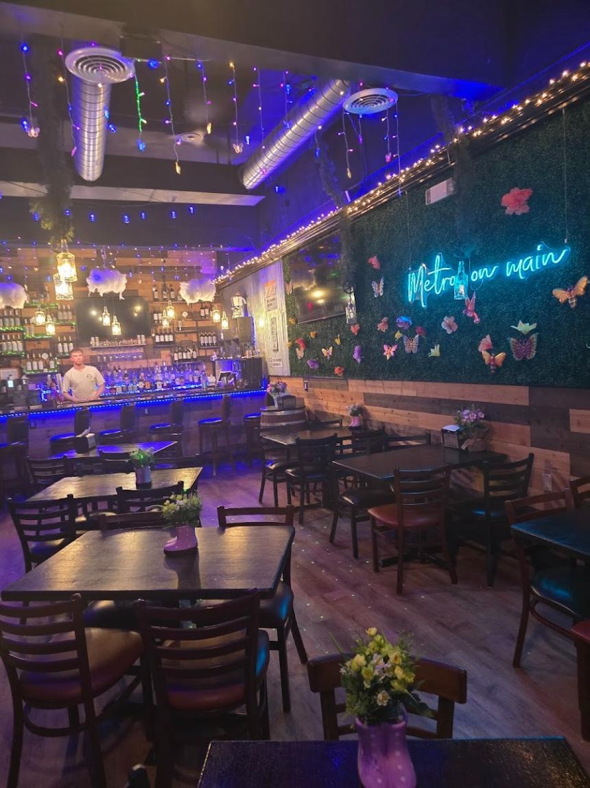 Cozy restaurant interior with wooden tables and chairs, colorful string lights hanging from the ceiling, a lit bar in the background, and a green wall decorated with butterflies and a glowing neon sign reading “Met her on main.